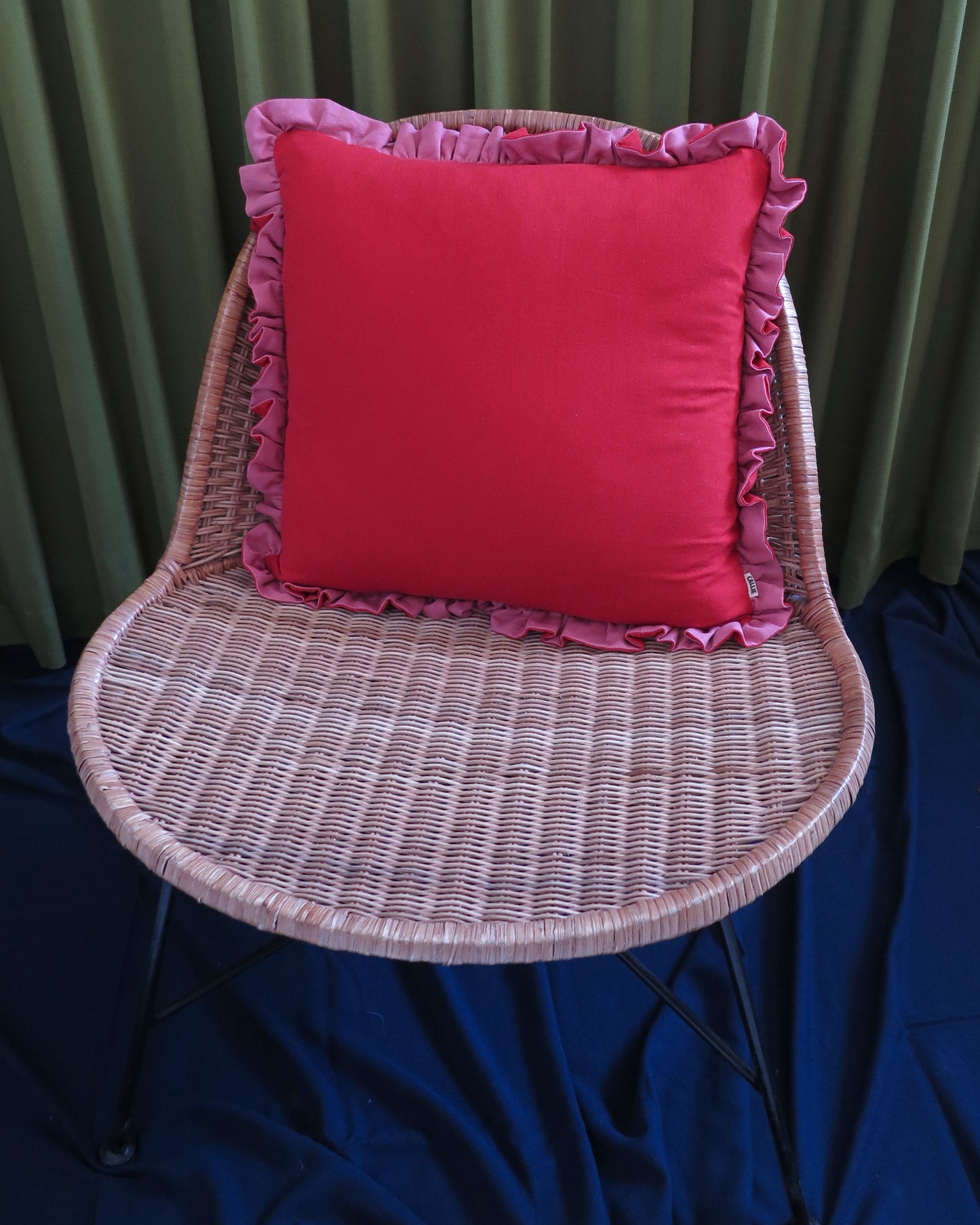 A bright red square throw pillow with pink ruffled trim, displayed on a woven wicker chair against a backdrop of green curtains and a blue fabric floor covering.