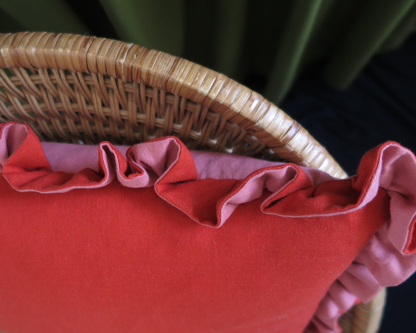 A close-up of a red cushion with pink ruffled trim, shown resting on a woven wicker chair.