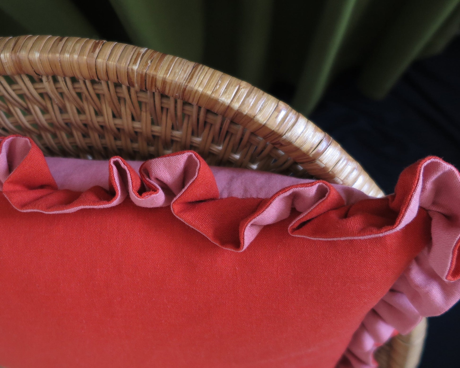 A close-up of a red cushion with pink ruffled trim, shown resting on a woven wicker chair.