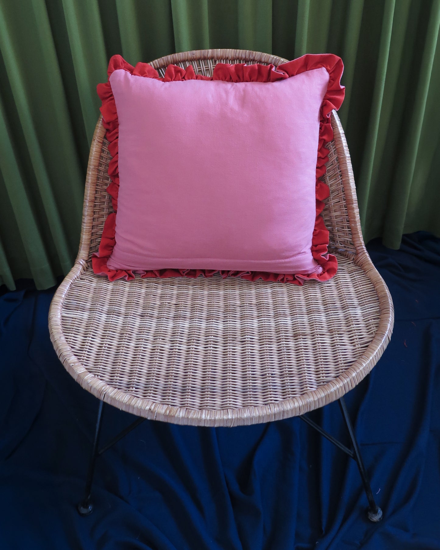 A pink square throw pillow with red ruffled trim, displayed on a woven wicker chair against a green curtain backdrop and dark blue floor covering.