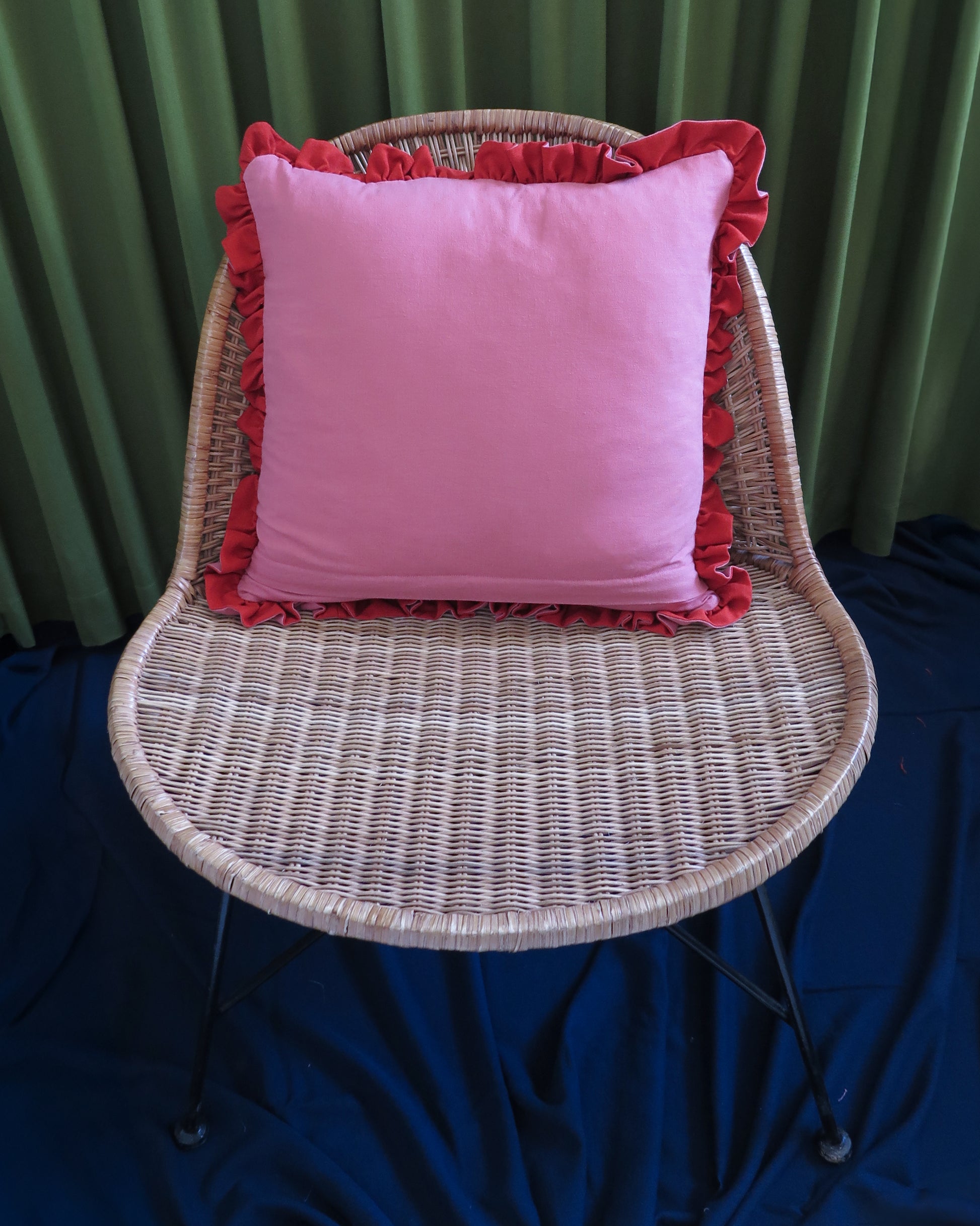 A pink square throw pillow with red ruffled trim, displayed on a woven wicker chair against a green curtain backdrop and dark blue floor covering.