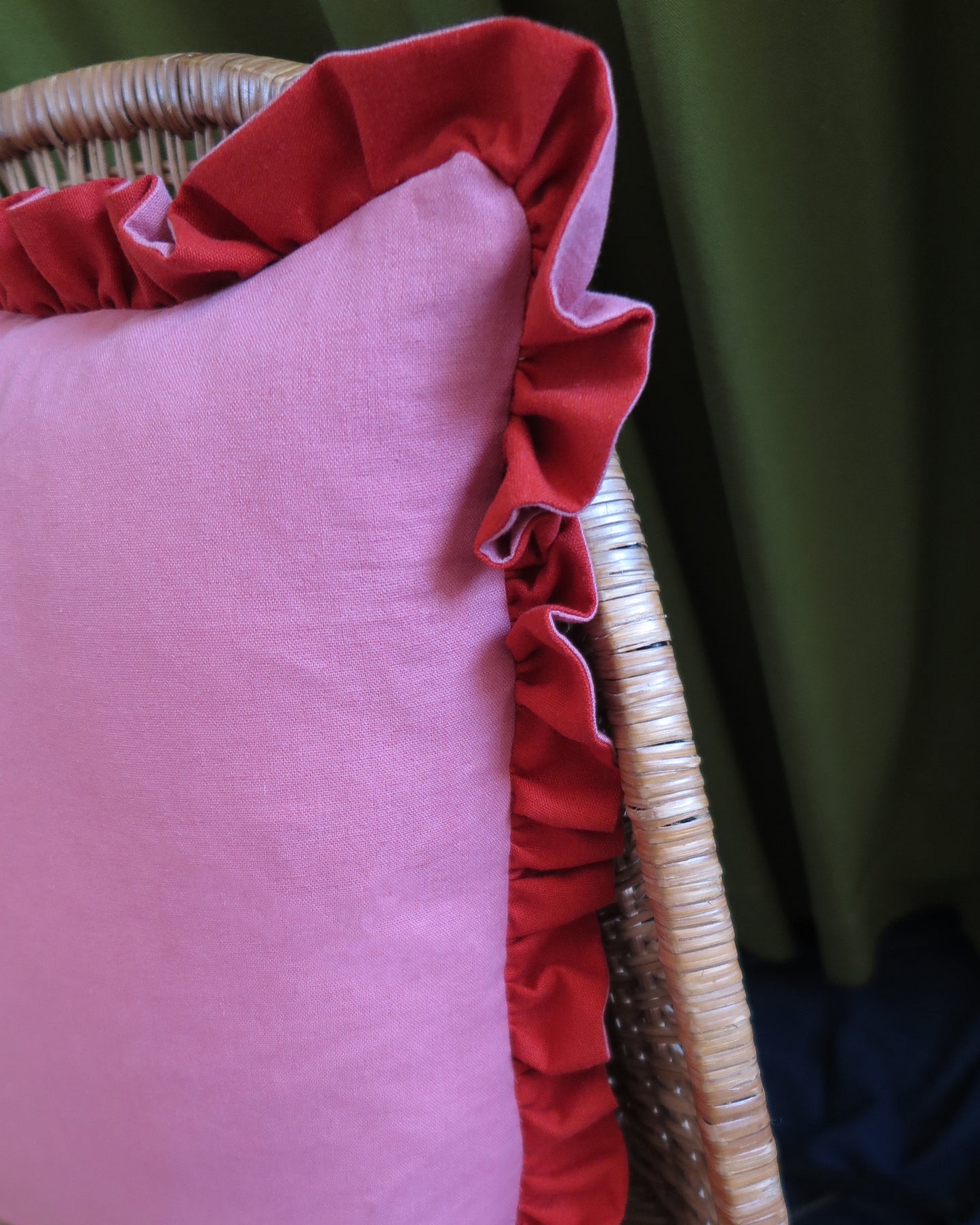 A close-up of a pink throw pillow with a red ruffled trim, resting on a wicker chair against a green curtain background.